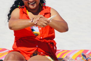 Mulher sorridente sentada à praia passando filtro solar no braço