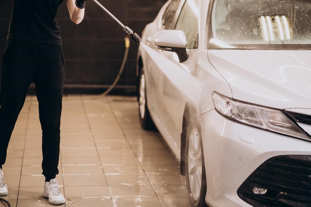 Pessoa lavando carro em casa, utilizando mangueira e produtos de limpeza para manter o veículo brilhante e bem cuidado.