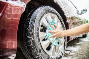 Pessoa lavando o carro em casa, aplicando sabão na roda do veículo. A prática de lavar o carro em casa é uma maneira econômica e eficiente de manter o automóvel limpo