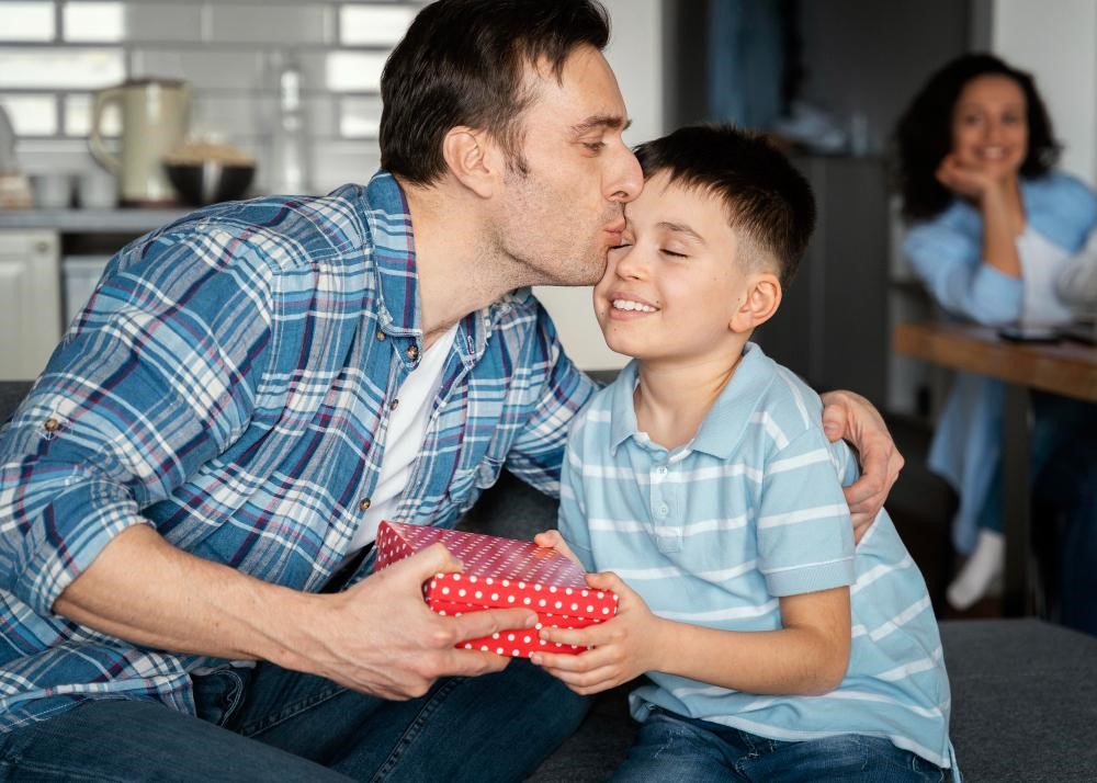 Um pai recebe um presente do filho em um momento de carinho e alegria. O menino, sorridente, entrega um embrulho colorido, enquanto o pai o beija na testa, simbolizando o amor e a conexão entre eles. Ao fundo, uma mulher observa a cena com um sorriso, refletindo a felicidade da família unida.