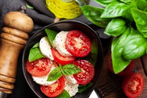 Uma salada colorida com fatias de tomate, queijo mozzarella e folhas de manjericão, dispostas em uma tigela escura. Ao fundo, ingredientes como pimenta-do-reino e limão complementam a cena.