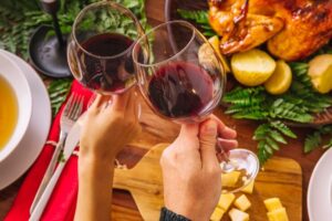 Duas taças de vinho tinto sendo brindadas, acompanhadas de um prato com carne assada e acompanhamentos. A composição destaca a harmonização de vinhos e carnes, perfeita para momentos de celebração.