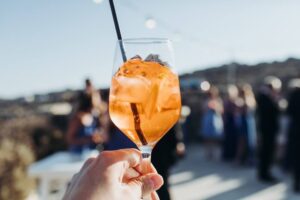 Foco em taça com Aperol tonic e ao fundo um ambiente festivo. 