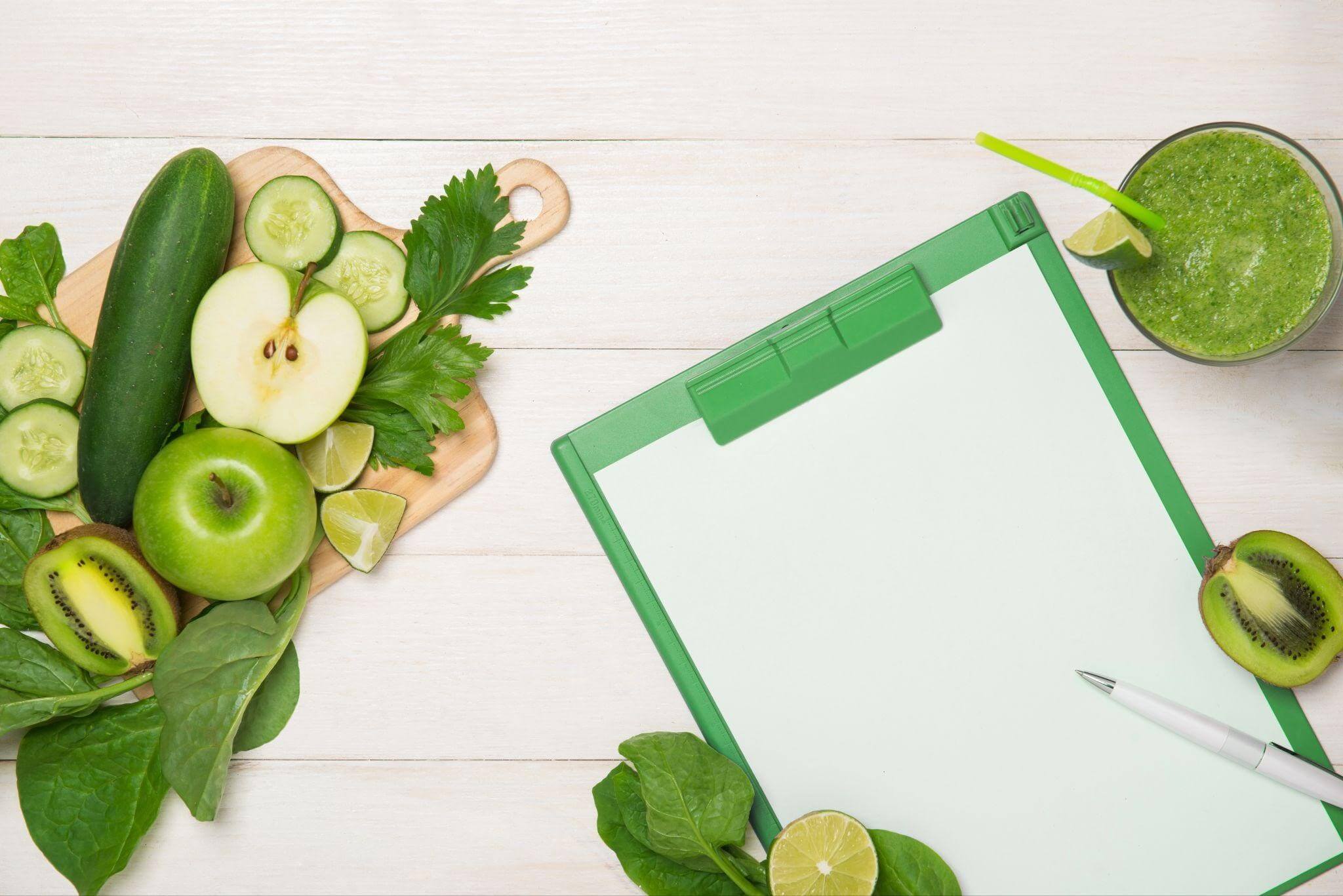 Imagem com diversos ingredientes frescos representando hábitos saudáveis, incluindo maçãs verdes, pepino, folhas verdes, limão e um smoothie saudável em um copo. Ao lado, um bloco de notas em um suporte verde, sugerindo planejamento e organização na adoção de hábitos saudáveis.