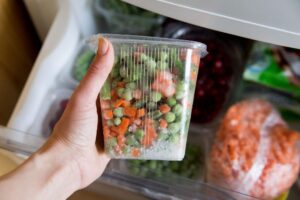  Mão segurando um recipiente transparente com legumes congelados e gelo, destacando a importância de identificar alimentos congelados e o correto armazenamento no freezer para garantir a segurança alimentar.