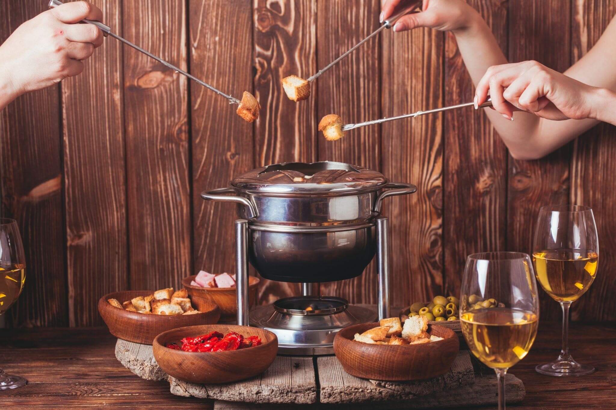 Mãos segurando espetos com pedaços de pão, mergulhando em uma panela de fondue em casa, cercada por pratos com acompanhamentos como azeitonas e tomates secos.