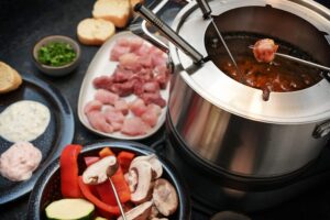 Um fondue com carnes e vegetais, com pedaços de carne crua sendo cozidos na panela quente, acompanhados por molhos e fatias de pão.