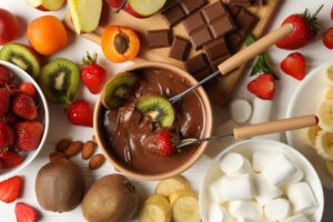 Uma variedade de frutas frescas como morangos, kiwis, e bananas, acompanhando uma tigela de fondue de chocolate, com espetos prontos para mergulhar as frutas.