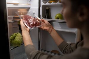Mulher colocando carne embrulhada em embalagem plástica na prateleira de um refrigerador, ilustrando os métodos corretos para o descongelamento seguro de alimentos, como a descongelação na geladeira.