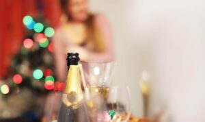 Close-up em uma garrafa de espumante aberta com rótulo dourado sobre uma mesa de celebração. Ao fundo, de forma desfocada, vê-se uma árvore de Natal com luzes coloridas acesas e uma pessoa com roupas em tons de rosa, criando uma atmosfera festiva e aconchegante.