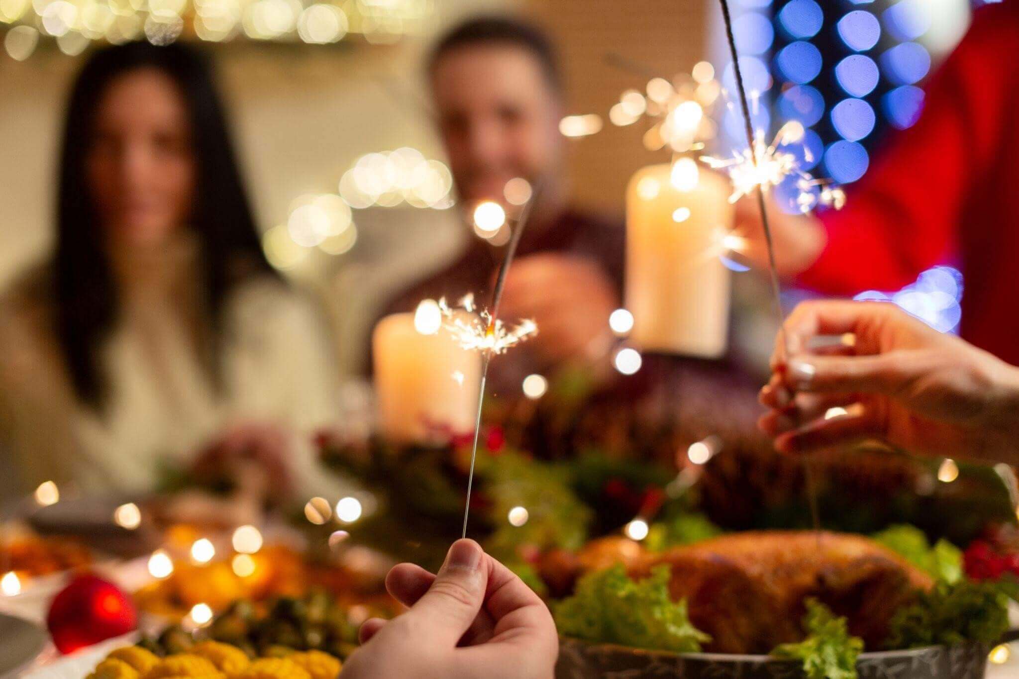 Foco seletivo em mãos segurando velas faíscas (sparklers) acesas sobre uma mesa de jantar natalina. O fundo está desfocado (efeito bokeh), mostrando silhuetas de pessoas sorrindo e luzes de Natal douradas e azuis.