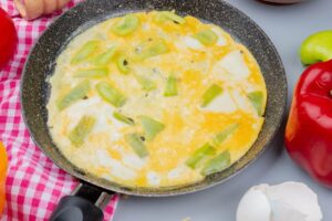 Foto em close de um omelete amarelinho sendo preparado em uma frigideira antiaderente, recheado com pedaços suculentos de pimentão verde. Ao lado, ingredientes frescos como pimentões inteiros e cascas de ovo, compondo uma das receitas para performance e recuperação muscular ricas em proteínas.