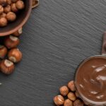 Vista superior de uma mesa de pedra escura com um pote de chocolate, avelãs naturais e pedaços de barra de chocolate para preparar receitas com creme de avelã.