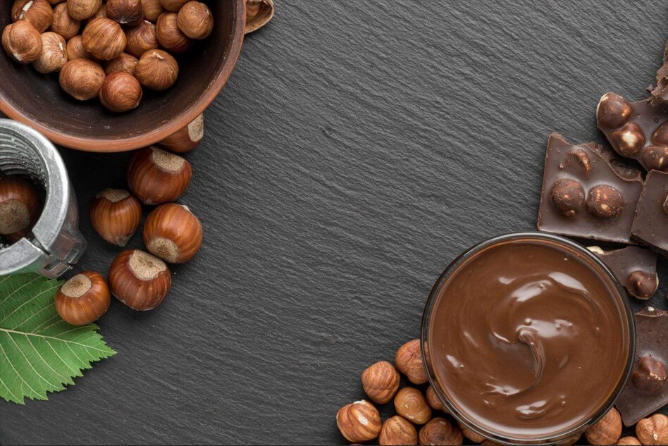 Vista superior de uma mesa de pedra escura com um pote de chocolate, avelãs naturais e pedaços de barra de chocolate para preparar receitas com creme de avelã.