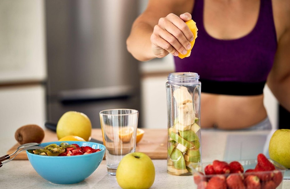 Uma mulher com roupas esportivas preparando um suco natural em uma cozinha iluminada. Ela espreme uma laranja sobre um liquidificador portátil que já contém frutas picadas, cercada por tigelas com morangos e kiwis, focando no preparo de receitas para performance e recuperação muscular.