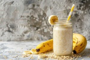 Um copo de vidro com um smoothie cremoso de banana e aveia, decorado com uma rodela de banana na borda e um canudo listrado. O copo está cercado por bananas frescas e flocos de aveia sobre uma superfície rústica, exemplificando receitas para performance e recuperação muscular ideais para o pós-treino.