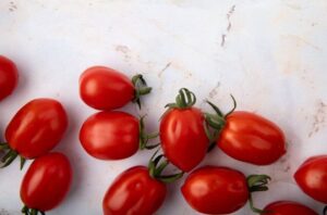 Diversos frutos de tomate italiano espalhados sobre uma superfície de mármore claro, mostrando seu formato oval característico e ramas verdes.