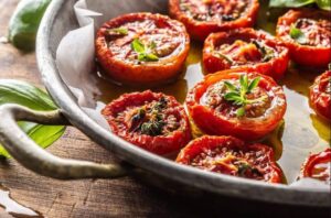 Close de fatias de tomate assadas em uma frigideira com azeite e ervas frescas, destacando a textura suculenta do fruto após o cozimento.