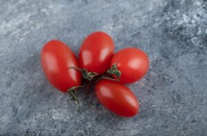 Quatro unidades de tomate tipo uva, com formato alongado e casca lisa, apresentados sobre um fundo cinza texturizado.