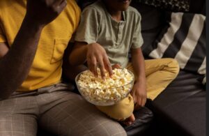 Foto em plano fechado de um pai e um filho sentados no sofá compartilhando uma tigela de vidro com pipoca branca. O foco está nas mãos pegando o lanche, ideal para quem busca momentos em família e receitas de pipoca simples.