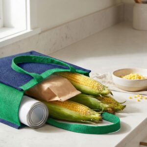 Uma sacola de compras reutilizável em tons de azul e verde sobre uma bancada de cozinha branca. Dentro da sacola, há espigas de milho frescas com palha, uma lata branca e um saco de papel pardo, sugerindo diferentes formas de aproveitar o milho desde o momento da compra. Ao fundo, um pequeno bowl contém grãos de milho cozidos. Imagem 2: Dicas para preparar a receita certa.jpg