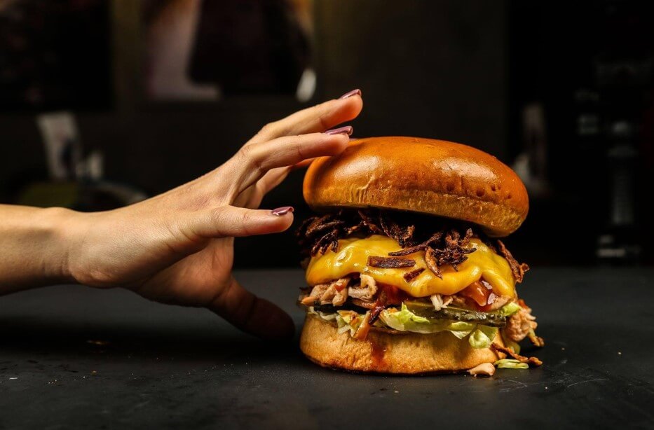 Close-up de um hambúrguer robusto montado em um pão para hambúrguer artesanal do tipo brioche, que possui uma superfície dourada e brilhante. O recheio conta com bastante queijo derretido, carne suína desfiada, cebola frita crocante, alface e picles. À esquerda, uma mão com unhas pintadas se aproxima para segurar o sanduíche, que está sobre um fundo escuro.