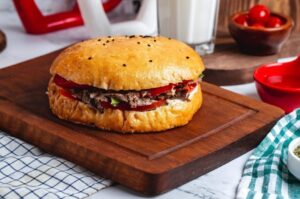 Um sanduíche montado em um pão para hambúrguer artesanal do tipo brioche, polvilhado com gergelim preto e branco na parte superior. O sanduíche está recheado com carne desfiada, fatias de tomate e molho, servido sobre uma tábua de madeira rústica ao lado de um copo de leite e um pote com tomates cereja.