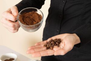 Pessoa segurando uma xícara de vidro com café moído em uma mão e grãos inteiros na outra, ilustrando a dúvida sobre qual o melhor recipiente e onde guardar o café moído ou em grão.