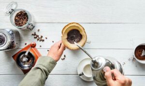 Vista aérea de uma mesa de madeira branca com potes de vidro herméticos, moedor manual e café sendo coado, ensinando onde guardar o café para manter o frescor durante o preparo artesanal.