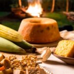 Mesa de doces e salgados típicos de festa junina arrumada ao ar livre com uma fogueira ao fundo. Em primeiro plano, há um cesto de palha com doce de leite, pé de moleque e paçoca, ao lado de milho verde cozido, pipoca derramada, bolo de fubá inteiro e em fatias, cachorro-quente e arroz doce. A imagem destaca a variedade de ingredientes para cardápio junino.