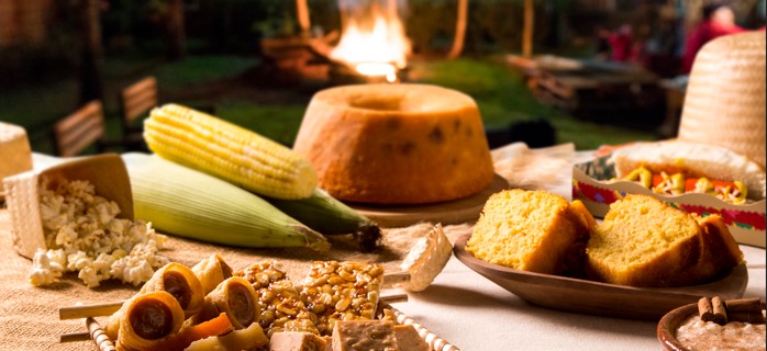 Mesa de doces e salgados típicos de festa junina arrumada ao ar livre com uma fogueira ao fundo. Em primeiro plano, há um cesto de palha com doce de leite, pé de moleque e paçoca, ao lado de milho verde cozido, pipoca derramada, bolo de fubá inteiro e em fatias, cachorro-quente e arroz doce. A imagem destaca a variedade de ingredientes para cardápio junino.