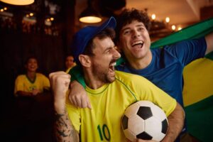 Duas pessoas com camisas da seleção brasileira comemoram alegremente em um ambiente interno. Um homem de boné azul e camisa amarela segura uma bola de futebol enquanto o outro o abraça por trás segurando a bandeira do Brasil. Ao fundo, outras pessoas assistem ao momento, ideal para ilustrar ideias sobre o que comprar para os jogos da Copa.